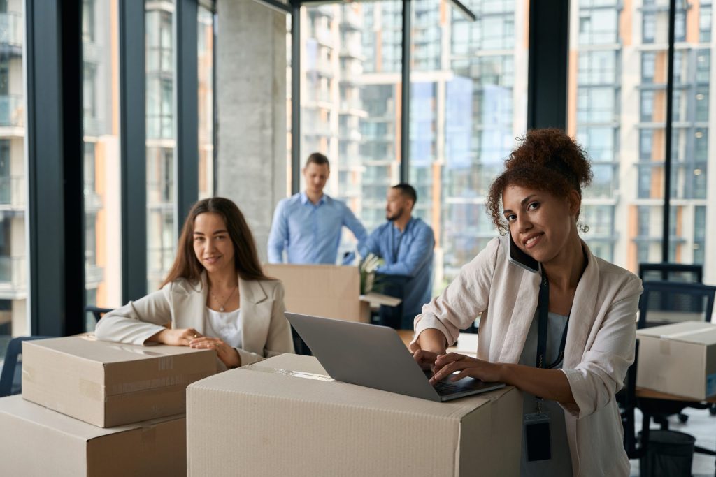 Joyful company worker working on laptop during phone conversation while her colleagues packing cardboard boxes